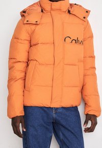 Pomarańczowa puchowa kurtka z wysokim kołnierzem, zamkiem błyskawicznym z przodu i czarnym logo. Posiada przeszywane panele oraz duże przednie kieszenie. W zestawie z niebieskimi jeansami.