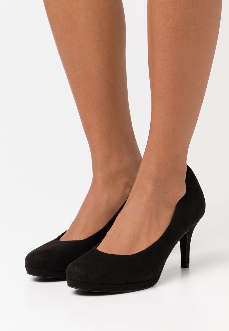 Pieds de femme portant des escarpins noirs en daim à bout fermé avec un talon fin, sur un fond blanc uni.