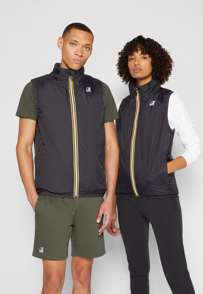 Sorte ermeløsevester med gule og hvite glidelåser, med sidelommer og en logo, bæres over grønne og svarte antrekk.