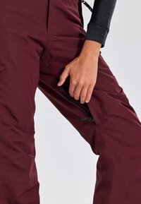 Pantaloni da neve color bordeaux con tasca con zip. Caratterizzati da un tessuto resistente, una vestibilità rilassata e bretelle regolabili per garantire comfort durante le attività invernali.
