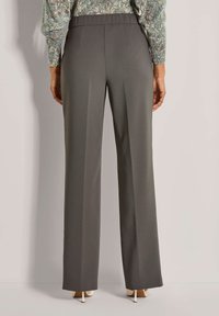 Persoon in grijze wijde broek en een bedrukte lange mouwtop, staand tegen een plain lichte achtergrond.