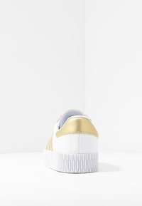 Sneaker bianco con tacco e dettagli laterali dorati, suola spessa e texturizzata bianca, mostrato da dietro su una superficie e sfondo bianchi.