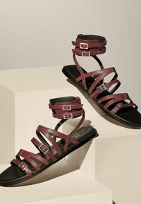 Mørkerød læder sandal med flere krydsede stropper og ankelspænder, som er sat på en sort sål. Teksturret, minimalistisk design med justerbare metaldele.