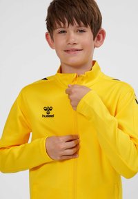 Junge mit kurzen braunen Haaren schließt den Reißverschluss einer leuchtend gelben Hummel-Sportjacke vor einfarbigem Hintergrund.