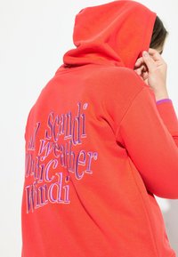 Sudadera roja con textura suave, que presenta un texto bordado multicolor en la parte posterior. Los puños son morados y la capucha es ajustable.
