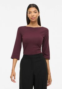 Bordō tonēts tops ar trim ceturtdaļas piedurknēm, apaļu kakla izgriezumu un mīkstu tekstūru, kombinēts ar piemērotām melnām biksēm.