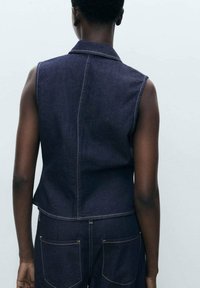 Mouwloze denim vest in donkerblauw, met achternaad en lichtgekleurde stiksels. Soepele textuur, cropped ontwerp, met ingesneden kraag.