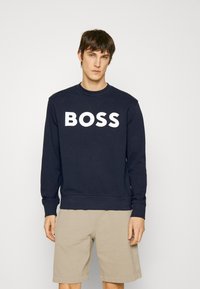 Sweatshirt azul-marinho com grande texto branco "BOSS" na frente, apresentando um clássico decote redondo e punhos e bainha canelados. Combinado com calções bege.
