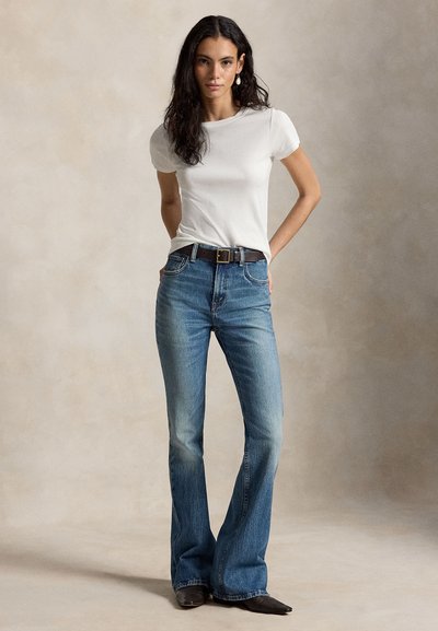Mujer de pie con las manos detrás de la espalda, vistiendo una camisa blanca de manga corta, jeans azules acampanados de talle alto, cinturón marrón y zapatos oscuros puntiagudos.