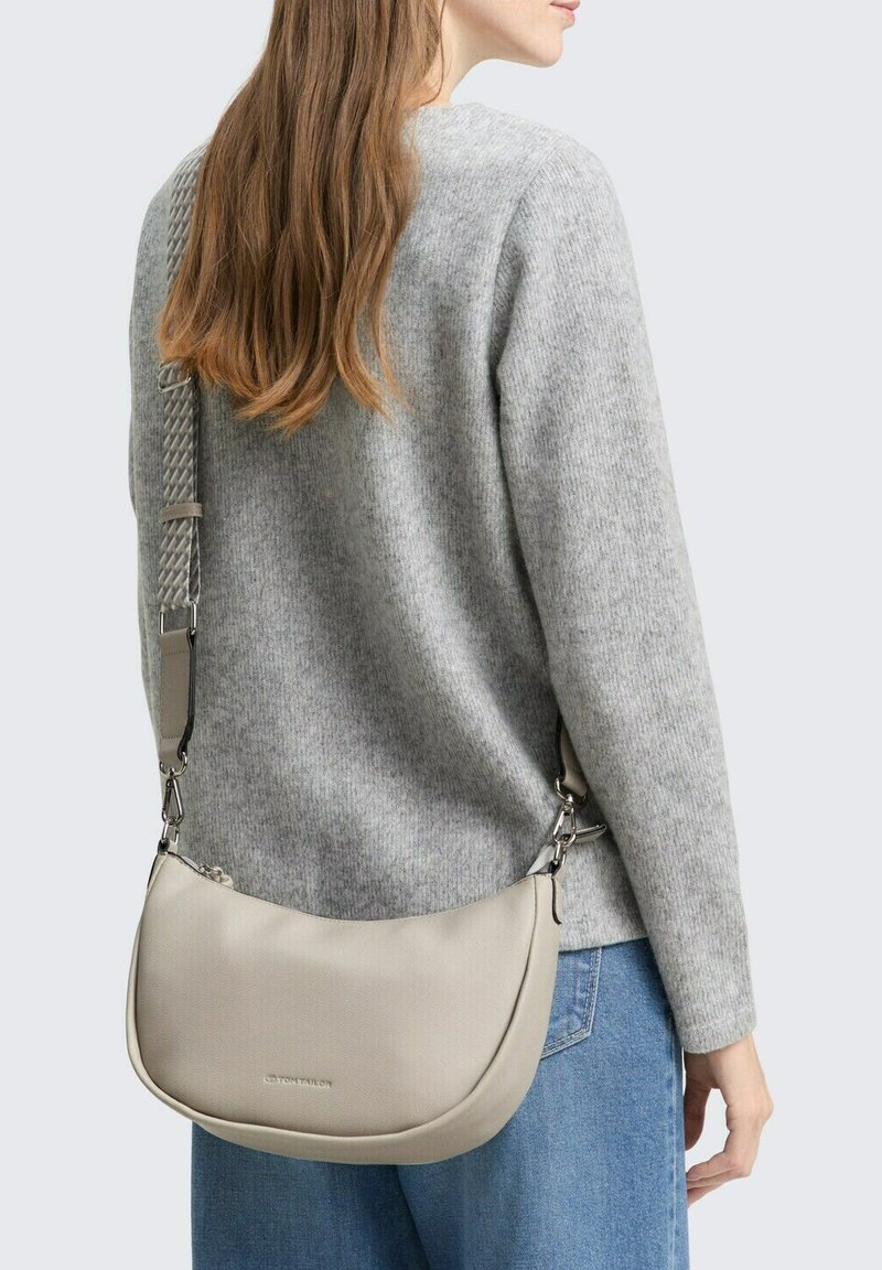 Borsa a tracolla beige con una texture liscia, forma semicircolare, accessori in argento e una tracolla intrecciata, indossata sopra un maglione grigio.