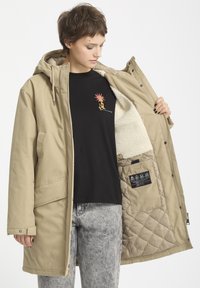 Beige parka dengan tudung, menampilkan lapisan lembut dan bertekstur. Bahagian dalamnya mempunyai corak berlapis dan terdapat label di dalam poket. Kemeja-T gelap di bawah.