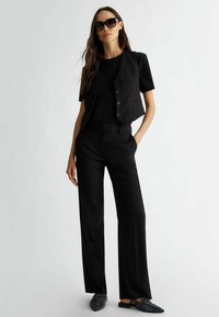 Gilet nero attillato con maniche corte, abbinato a pantaloni dritti con sottili motivi a pois e eleganti scarpe slip-on nere decorate.
