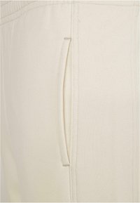 Pantaloni de trening bej deschis dintr-un material texturat, cu un buzunar lateral și detalii cusute de-a lungul cusăturilor.