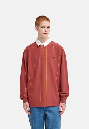 Jong persoon met kort rood haar draagt een rood rugbyshirt met dunne gele strepen, witte kraag en blauwe jeans, staand tegen een effen achtergrond.