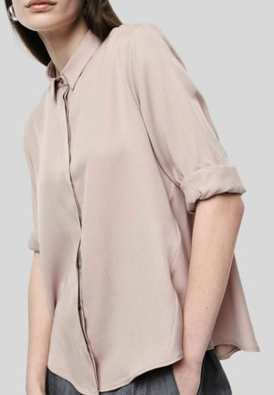 Donna che indossa una camicia rosa pallido a maniche lunghe, con le maniche arrotolate e la mano in tasca su sfondo grigio.