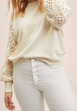 Femme portant un pull crème avec des manches en dentelle au crochet et un pantalon beige clair taille haute, debout devant un fond neutre.