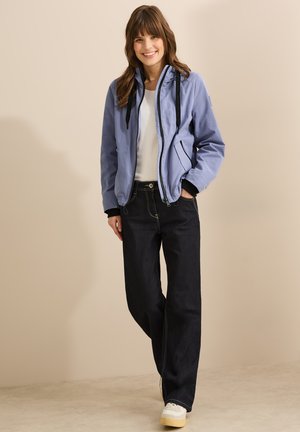 Light blue bomber jacket with black trim, zippered front, and drawstring hood, paired with dark jeans and white sneakers. Smooth texture.