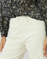 Pantalon en velours côtelé blanc avec des côtes verticales, présentant une fermeture à bouton à l'avant, des poches latérales et une coupe ajustée. Associé à un blouse noire à fleurs.