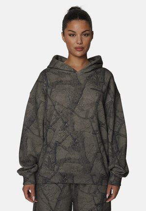 Vrouw die een donkergrijze en zwarte camouflageprint hoodie met bijpassende broek draagt, staand tegen een effen achtergrond.