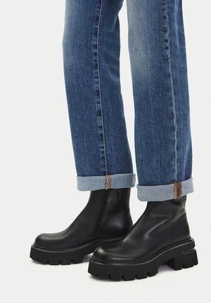 Schwarze Leder-Stiefeletten mit klobiger Sohle, seitlichem Reißverschluss und abgerundetem Zehenbereich, kombiniert mit hochgekrempelten blauen Jeans.