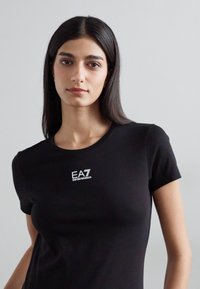 Melns pieguļošs T-krekls, kas izgatavots no gludas auduma. Priekšpusē neliels balts EA7 logotips. Īsās piedurknes un klasisks apaļais kakls.