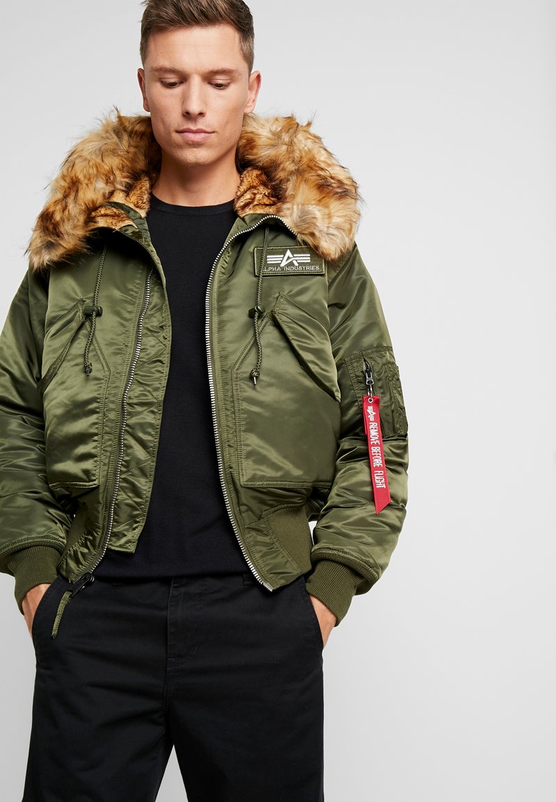 Maslinasto zeleni bomber jakna s kapuljačom od umjetnog krzna, zatvaranjem na zatvarač i više džepova, s crvenom privjeskom na ključeve i oznakom s logotipom.