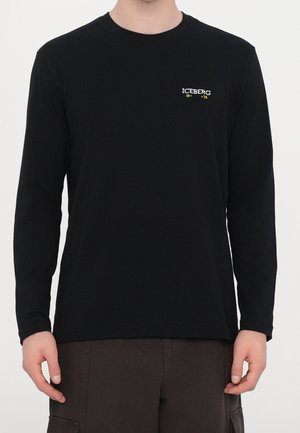 Homme portant une chemise noire à manches longues avec le logo "ICEBERG 1974" sur la poitrine gauche et un pantalon marron foncé avec poches cargo.