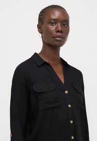 Blouse noire à manches longues en tissu léger, avec un col en V, deux poches poitrine et des accents de boutons sur le devant et les manches.