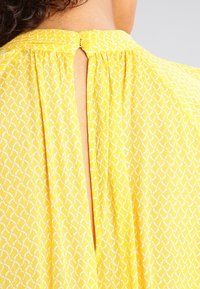 Blouse jaune avec un motif texturé de tourbillons blancs, fermeture à boutons au dos et une fente à l'encolure. Tissu doux et léger.