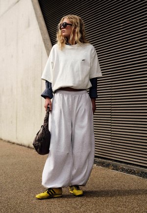 Woman with wavy blonde hair wears oversized white shirt and pants, yellow sneakers, sunglasses, and holds a brown handbag standing by a modern building.