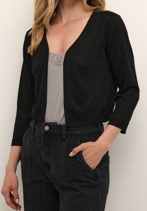 Femme portant un cardigan noir court sur un haut gris à bordure en dentelle, associé à un jean noir taille haute, main dans la poche.