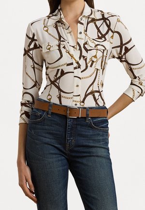 Vrouw draagt een wit overhemd met knopen met een bruine ketting- en riemprint, ingestopt in donkerblauwe spijkerbroek met een bruine leren riem.