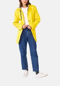 Imperméable jaune à capuche, avec des boutons-pression et une doublure intérieure rayée contrastante, porté sur un pantalon en denim bleu et des baskets blanches.