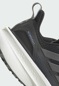 Chaussure de sport noire et grise avec une tige texturée, marquage ULTRA BOOST et une semelle intermédiaire blanche et nervurée. La semelle extérieure en caoutchouc durable présente des rainures.