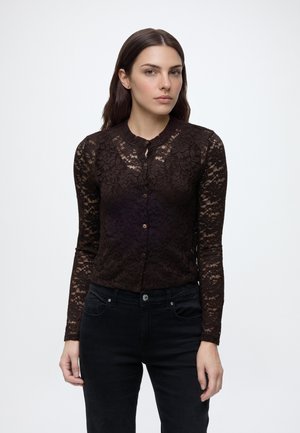 Femme debout portant un top noir en dentelle à manches longues avec des boutons et un pantalon noir sur un fond clair uni.