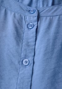 Camicetta azzurro chiaro con una texture liscia, dotata di bottoni in plastica trasparente e chiusura a bottoni. Colletto piatto e dettagli delle cuciture visibili.