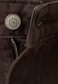 Gros plan d'un jean en tissu marron foncé montrant un bouton en métal avec des lettres et une fermeture éclair ouverte en dessous.