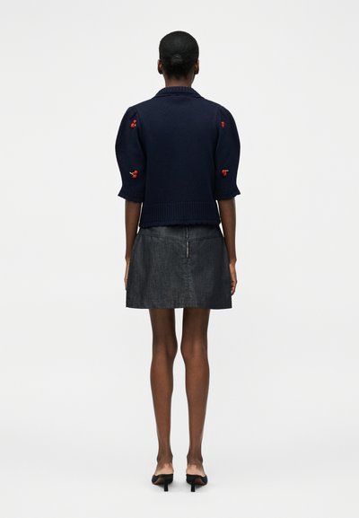 Γυναίκα με σκούρο δέρμα και μαλλιά σε κότσο φοράει ένα μπλε σκούρο κοντομάνικο πουλόβερ με κόκκινη κεντημένη κερασιά και μια μαύρη δερμάτινη μίνι φούστα με φερμουάρ στο πίσω μέρος.