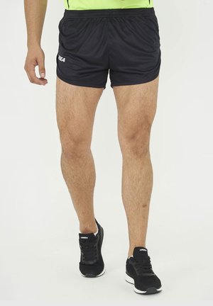 Nogi osoby noszącej czarne spodenki sportowe i czarne buty do biegania z białymi podeszwami, stojącej na białym tle.