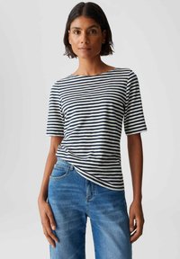 Femme aux cheveux courts et foncés portant une chemise à rayures noir et blanc à manches courtes rentrée dans un jean en denim bleu, sur fond uni.