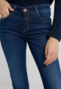 Jeans de mezclilla azul oscuro con un diseño ajustado, estilo de cinco bolsillos y detalle de costura naranja en la cintura y los bolsillos.