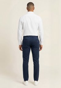 Mand stående med ryggen til, iført en hvid langærmet skjorte, mørkeblå bukser og hvide sneakers mod en neutral baggrund.