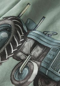 Zaļš audums ar detalizētu grafiku par vintage traktoru, ar lielām melnām riepām, metāla akcentiem un sarežģītiem dizaina elementiem.