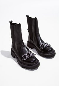 Bottines en cuir noir avec une semelle épaisse, dotées de détails en chaîne argentée sur le devant et de tirettes à l'arrière pour un enfilage facile.