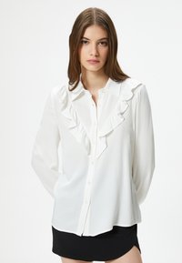 Camicia bianca con bottoni, con dettagli arricciati lungo la parte anteriore e il colletto, realizzata in un tessuto morbido, maniche lunghe e orlo curvo.
