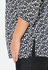 Blusa nera floreale con motivi bianchi, caratterizzata da maniche corte, uno spacco laterale e un tessuto leggero e testurizzato. Lo sfondo mostra denim nero.