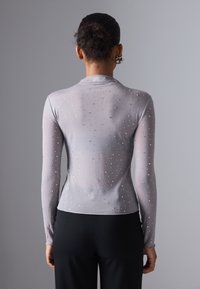 Svijetlosiva bluzica dugih rukava s prozirnom teksturom, ukrašena raspršenim rhinestones kamenčićima i visokim ovratnikom, u kombinaciji s crnim hlačama.