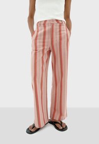 Gestreepte wijde broek in lichtroze en koraal met verticale lijnen, voorzien van een knoopsluiting en zijzakken. Gemaakt om te dragen met zwarte slippers.