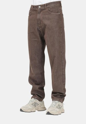 Jeans marroni a gamba dritta indossati con sneakers New Balance bianche e grigie su una persona in piedi contro uno sfondo semplice.