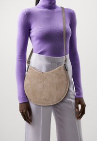 Beżowa zamszowa torebka crossbody w kształcie półksiężyca. Posiada zamek błyskawiczny oraz srebrne okucia. Nosi ją na lekko fioletowym golfie i jasnych spodniach.